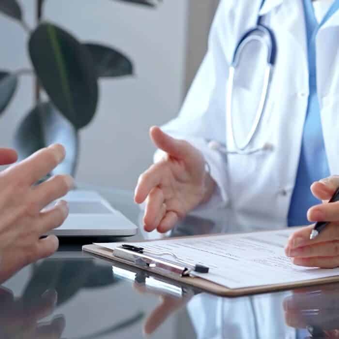 Doctor woman wearing white medical coat is consulting patient in clinic office while both are sitting at the glass desk. Physician is listening and making notes. Medicine concept.